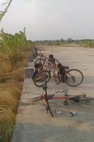 The video shows a group of children gathered around bicycles on a concrete surface next to a grassy area with banana trees. Two boys are actively working on a bicycle, with one appearing to be looking at the chain or gears. Another boy is standing nearby, holding a yellow object, possibly a banana or fruit. In the foreground, another bicycle is lying on its side, along with a pair of flip-flops. The sky is bright, suggesting daylight. The overall scene depicts children engaged in an activity with bicycles outdoors.
