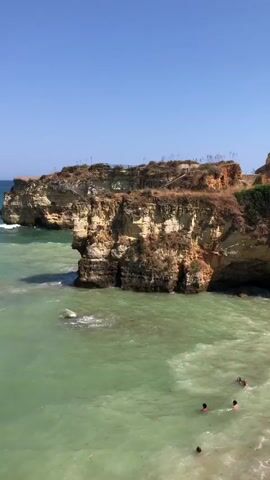 The video displays a scenic coastal view with a bright blue sky and clear water. Rocky cliffs are visible on the left side of the frame, with vegetation growing on top. The water is a vibrant turquoise green, with some areas appearing lighter due to shallow depths or sunlight. Small waves are gently lapping against the rocks. In the distance, there are faint outlines of land and possibly buildings on the horizon. A few small sailboats can be seen dotted across the sea. Towards the bottom right of the frame, several people are visible in the water, suggesting it is a beach or swimming area. The video pans slowly across the landscape, offering different perspectives of the coastline and the sea.