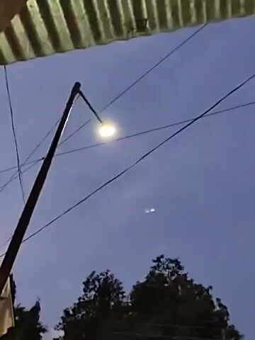 The video shows an outdoor scene at dusk. A street light is illuminated against a darkening blue sky. In the lower portion of the frame, trees are visible. The camera then pans to the right, revealing a residential area. Houses with lights on can be seen, along with parked vehicles, including a car and a motorcycle. The overall impression is of a quiet suburban or rural street as night begins to fall.