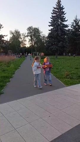Two young girls are walking on a paved path in a park-like setting. They are both carrying boxes of pizza. The girl on the left is wearing a white hooded sweatshirt with the word "Creative" visible on it, and light-wash ripped jeans. The girl on the right is wearing a purple jacket over a pink shirt and baggy jeans with patches. They appear to be walking towards the viewer.

The setting is a park with green grass, trees, and a pathway. In the background, there are other people walking and some playground equipment. The time of day appears to be late afternoon or early evening, with the sun casting a warm glow and long shadows. The sky is a pale blue, suggesting fair weather. The location is identified as Chernogolovka, Russia. The overall mood seems casual and lighthearted, with the girls engaged in a simple outdoor activity.