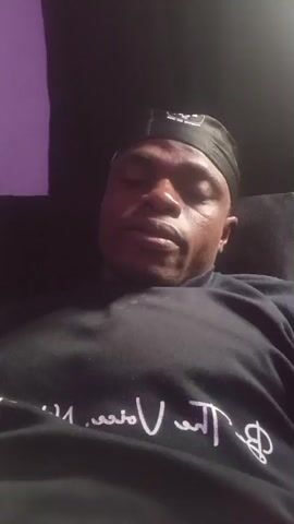 A man wearing a black baseball cap and a black t-shirt with white lettering is looking at the camera. The man is seated in a dimly lit room. To his left, a section of a wall is lit with a purple light. The man appears to be speaking or reacting to something off-camera.