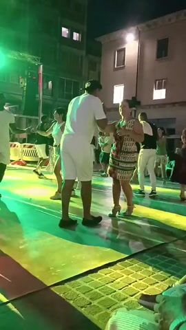 The video shows a night scene with a group of people dancing. The primary focus is on a man in a white t-shirt and white shorts dancing with a woman wearing a patterned dress. They are in the foreground and appear to be the main couple dancing. The lighting is predominantly green and blue, creating a festive atmosphere. In the background, other people are also dancing and there are buildings with illuminated windows. The ground appears to be paved. A stage with lighting equipment is visible on the left side of the frame. The overall impression is of an outdoor party or festival.