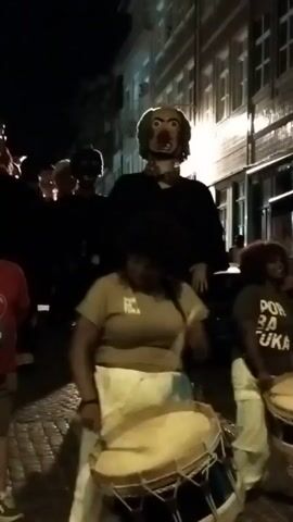 A group of people are in the street at night. In the background, large figures of exaggerated, grotesque faces stand. In the foreground, two women are playing large drums. One woman is wearing a tan t-shirt and white pants. The other woman is wearing a dark t-shirt with white text and light-colored pants. The street appears to be paved. Buildings are visible along the side of the street.