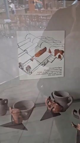 The video pans across a display case containing ancient pottery fragments and a framed illustration. The illustration depicts a cross-section of a tiled roof, showing the arrangement of terracotta tiles and coping stones. Text below the illustration, in Italian, describes the roof covering of a temple. The pottery fragments in the display case appear to be shards of various vessels, including bowls and possibly a jug. The environment suggests a museum or exhibition setting, with a muted background and lighting focused on the display.