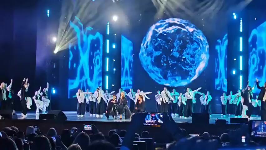 The video shows a stage performance with multiple dancers and a singer. The stage is lit with blue and white lights, and a large spherical screen displays a blue, watery image. On the stage, a group of dancers in white shirts and black pants perform synchronized movements. In the center of the stage, a woman in a wheelchair sings into a microphone. She is wearing a dark outfit with a feathered skirt. Two men in black outfits are kneeling on either side of her. The audience is visible in the foreground, with many people holding up their phones to record the performance. The overall atmosphere appears to be a lively and energetic concert or show.