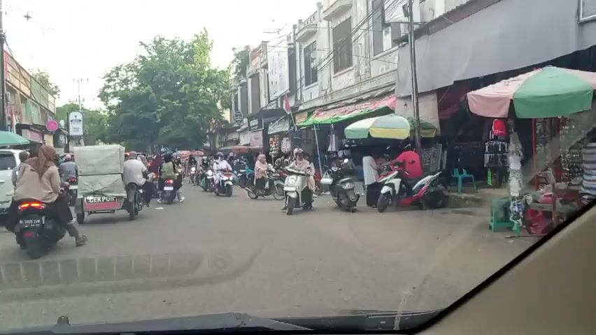 The video shows a busy street in what appears to be a Southeast Asian country. The view is from inside a vehicle, looking out onto a street crowded with motorcycles. Most of the riders are wearing helmets, and some are carrying passengers. The street is lined with shops and stalls, many of which have awnings or umbrellas providing shade. The buildings are mostly two or three stories tall, and some have ornate facades. There's a white car to the left, and utility poles and wires are visible overhead. The overall scene is one of bustling activity, typical of a commercial street in the region.