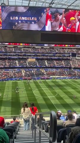A high-angle shot shows a soccer stadium during a match. The field is green with white markings, and several players in dark uniforms are scattered across it. The stands are filled with spectators, many wearing red. A large screen at the top displays "LOS ANGELES" in large letters with an American flag design behind it. In the foreground, seated spectators face the field. Two women in the middle ground stand and look towards the field. The stadium appears to be open-air.
