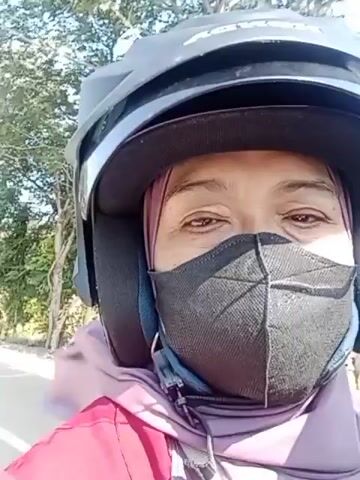 A close-up, first-person perspective video shows a person wearing a black helmet and a black face mask. They are also wearing a pink headscarf and a red jacket. The person is looking at the camera and smiling. In the background, there are trees and a building. The video appears to have been filmed outdoors on a sunny day. The person is riding a motorcycle or a similar vehicle.