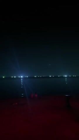 A night shot shows a group of people on a beach next to the sea. They are standing and sitting around tables and chairs illuminated by red lighting. A tree with lights is also visible. In the distance, there are lights on the water.