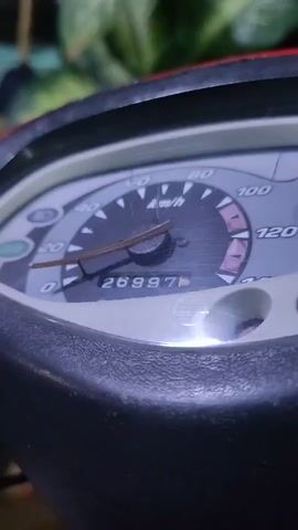 The video shows a close-up of a motorcycle's odometer. The odometer displays the mileage as 26997. The speedometer needle is pointing slightly above the 0 mark. The background is blurred but appears to be foliage.