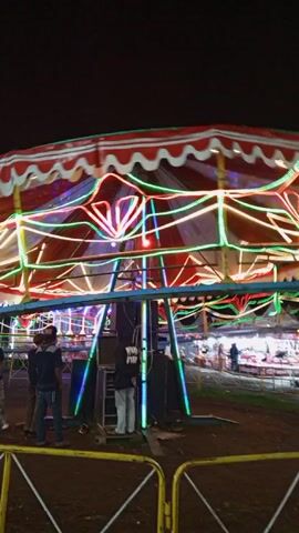 The video shows a carnival ride at night. The ride has a red-and-white striped canopy, with colorful string lights underneath. The ride is spinning. Three people stand near the ride, appearing to be watching it. There are speakers next to them. Other people are visible in the background, as are tents or booths, also lit up. The ground is dark, probably dirt or grass.