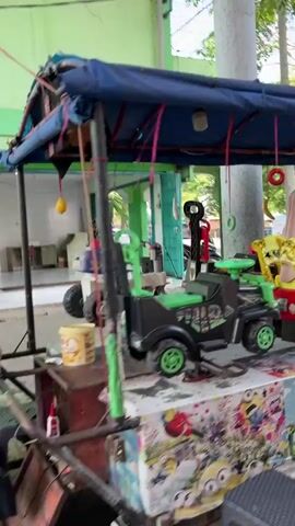 A group of people are gathered around a small amusement ride with colorful plastic cars. A child is seated in a red ride-on car, covered with a light-colored hijab and wearing a blue shirt. Another child, possibly in a yellow seat, is visible in the background. A woman with a brown hijab and green shirt stands to the left, observing the ride. To the right, another woman in a pink patterned shirt and dark pants is holding onto the ride and interacting with a child in a blue car. The ride is decorated with various cartoon characters and colorful graphics. The environment appears to be outdoors, under a blue canopy supported by metal poles, with a light green wall in the background. The ground is covered with what looks like a patterned mat or tarp.