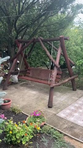 A wooden swing set with a bench is situated on a concrete patio. The swing set is made of natural wood logs, forming an A-frame structure. The bench has a patterned cushion. In the foreground, there is a garden bed with various plants and flowers, including some blue spruce branches on the left. The background features trees and foliage, with what appears to be a house or building on the far left. The lighting suggests an overcast day. The video shows subtle movements, likely due to wind or the natural settling of the swing.