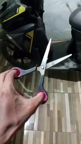 A hand is holding a pair of open scissors in the foreground. The scissors have pink grips. In the background, a black and yellow scooter is parked on a tiled floor.