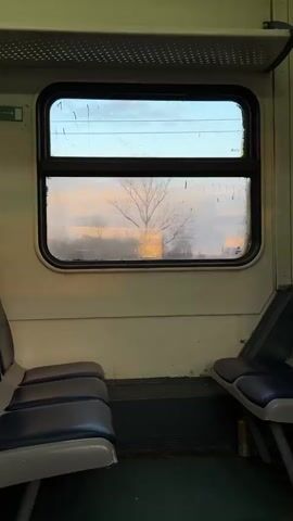 A vertical indoor shot shows the interior of a train car, likely during sunset. Two sets of blue seats are visible, facing each other, with a window in between. The window is slightly foggy, with condensation droplets, offering a view of the outside. Trees are visible, as well as a building in the distance, all cast in the warm glow of the sunset. The train car's interior is lit with a soft, warm light.