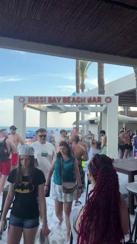 The video captures a bustling daytime scene at the "NISSI BAY BEACH BAR" in Ayia Napa, Cyprus. Numerous people are gathered outdoors, enjoying what appears to be a foam party. White foam is actively being sprayed from structures within the bar area, covering the ground and patrons.

Many individuals, dressed in swimwear or casual beach attire, are walking through, standing in, or around the foam. Some are dancing, while others are mingling or holding drinks at small tables. The atmosphere is lively and festive.

In the background, beyond the main party area, palm trees and resort buildings are visible under a clear, sunny sky. The sea can be faintly seen in the distance, confirming the beachside location. The prominent sign for "NISSI BAY BEACH BAR" is clearly displayed in red letters, with "nb" logos on either side. The overall impression is one of a vibrant summer beach party.