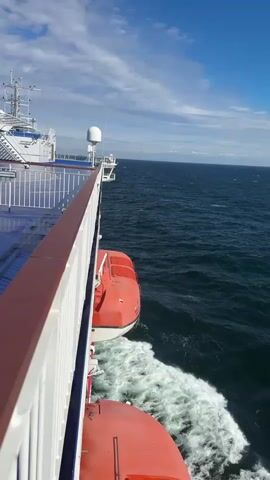 This video captures a view from the deck of a large ferry moving across a body of water. The camera is positioned along the railing, looking out towards the open sea.

**Subjects:**
*   **Objects:** The prominent objects include the ferry itself with its white railings, lifeboats (bright orange), and various antennas and structures on the upper decks. The sea is a significant element, with waves and white foam trailing behind the vessel.
*   **Scene/Setting:** The scene is the open sea, with a clear blue sky and scattered clouds. The ferry is in motion, indicated by the wake it leaves.
*   **People:** In the distance on the upper deck of the ferry, a few individuals can be seen seated, enjoying the view or traveling.

**Activity or Event:**
The primary activity is the ferry's passage across the sea. The movement creates a constant churn of water and foam in its wake.

**Notable Details:**
*   **Time of Day:** The bright sunlight and blue sky suggest it is daytime, likely late morning or afternoon.
*   **Weather:** The weather appears pleasant with a clear sky and a moderate amount of wave activity on the sea, indicating a breezy but not stormy day.
*   **Location Cues:** While the exact location is unknown, the open sea and the presence of a large ferry suggest a commercial shipping route or a passenger ferry service.
*   **Emotions:** The imagery evokes a sense of travel, freedom, and the vastness of the ocean. The clear weather adds to a feeling of calm and optimism.

**Visible Text:**
The numeral "3" is visible on one of the orange lifeboats.