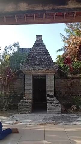 A video shows an outdoor scene with a stone structure at the center. The structure is a brick building with a pyramid-shaped roof. It has an open doorway with steps leading up to it. A broom is leaning against the right side of the doorway. There are plants and trees around the building, and the sky is visible above. A person's legs and feet are visible in the bottom left corner of the frame, suggesting they are sitting or kneeling. The scene appears to be in a sunny environment.