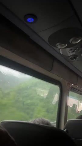 The camera is positioned inside a bus, looking out the window. Rain streaks across the windowpane, obscuring the view of lush green hills outside. A red and white sign is partially visible through the rain-streaked window. Inside the bus, the gray interior is visible, including the overhead console with multiple round vents and a blue light. The back of a passenger's gray head is visible in the foreground. The camera is relatively still.