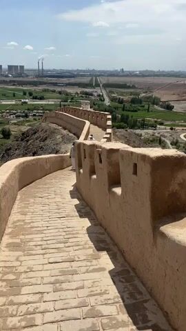 The video shows a perspective from on top of a tan-colored ancient wall with square crenellations. The wall has a brick pathway. A person in traditional clothing is visible walking up the pathway on the left. The background shows a valley with greenery, a road, and industrial buildings with smokestacks in the distance. The sky is blue with white clouds. The camera pans to the right, showing more of the wall and the mountainous landscape in the distance.