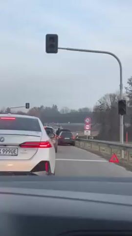 The video shows a view from inside a car stopped at a red traffic light.  Ahead, several cars are also waiting. A white BMW with the license plate "HN K 9990E" is directly in front of the camera car. Further ahead, a dark-colored car has its trunk open, and a person is visible near it. To the right of the road, there are speed limit signs indicating 70 km/h. An emergency triangle is placed on the side of the road.  The environment appears to be a rural area with trees visible in the background. The sky is overcast.