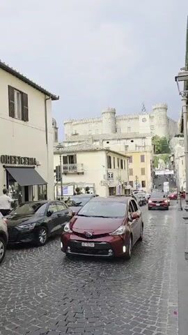 The media file captures a street scene in Bracciano, Italy, likely during the daytime. A red hybrid car is prominently featured in the foreground, moving down a cobblestone street. In the background, a large, pale building with castle-like turrets dominates the skyline, suggesting a historic structure such as a palazzo or fortress.

Several other vehicles, including dark sedans and a red car further down the street, are also present, indicating moderate traffic. Pedestrians are visible on the sidewalks, one of whom appears to be near a shop with signage that reads "FIOREFICERIA". The architecture of the buildings lining the street is typical of Italian towns, with cream-colored facades and shuttered windows. A lamppost is visible on the right side of the frame. The sky appears overcast, suggesting cloudy weather. The overall atmosphere seems to be that of a quiet, historic town with some vehicular activity.