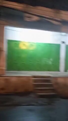 The video shows a blurry, handheld shot of a building facade at night. The building appears to be an urban commercial space with multiple storefronts or entrances. The facade is lit by artificial lights, creating a mix of shadows and illuminated areas. Several sections of the facade are covered with green or tan colored shutters or panels, some of which appear to be roll-up doors. There are also visible stairs leading up to some of the entrances. The overall impression is of a street-level commercial area that is closed for the night.