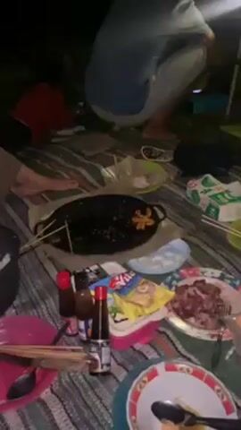 A group of four people are sitting on a striped blanket on the ground in the dark. There is a spread of food between them. In the center of the frame are plates with food remnants, a skillet with skewers, small bottles of condiments, and packaged snacks. One person in a pink shirt is sitting and looking at a phone. Another person in a red shirt is lying down, also looking at a phone. A third person in a blue shirt is squatting and looking at something out of frame. A fourth person is partially visible on the left side of the frame. There is a pot of rice next to them. The environment appears to be outdoors at night.