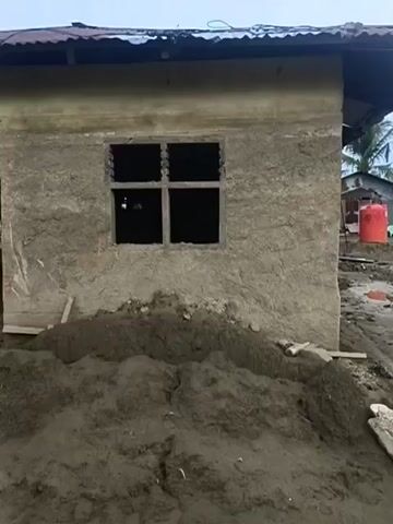 The video shows the exterior of a building under construction, possibly made of mud or earth. The walls are rough and unplastered, with a window opening that has wooden slats. There is a pile of dirt or sand in front of the building, and the ground is muddy. The roof appears to be made of corrugated metal. To the left of the main building, another similar structure is visible, also appearing to be under construction. The overall impression is of a rural or developing area.