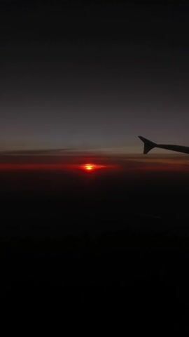 The scene is captured from the inside of an airplane, as evidenced by the view of a plane wing in the upper right corner. It is either sunrise or sunset, with a bright red sun visible on the horizon. The overall tone is dark, as the foreground and the upper sky are dimly lit. The sky is a mixture of grays and hints of orange near the horizon. The scene is stable, suggesting the plane is in stable flight during the recording.