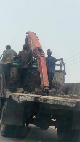 Three men stand in the back of a dirty, pale green dump truck with a rusty orange crane attached. One man wears a yellow and green shirt, another a dark shirt and cap, and the third a black and blue striped shirt and a black helmet. The crane is positioned over the truck bed, which contains dirt and debris. The background is a bright, overcast sky. The truck is parked on the side of a road.