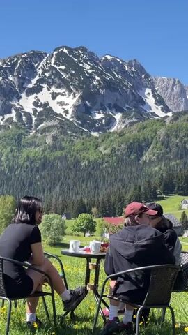 Four people are seated around a small round table outdoors on a sunny day. Two white mugs and a platter with food are on the table. Behind them is a mountain range, with snow on the peaks. In front of the mountains is a landscape of trees and grass. The people are chatting. The person on the left wears a black dress and sneakers. The next person wears a red baseball cap and a black hoodie. The third wears a black baseball cap and sunglasses. The fourth person is mostly hidden from view.