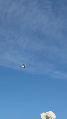 The video captures a clear blue sky with scattered white clouds. A white passenger airplane is seen flying horizontally across the frame from left to right. The bottom of the frame shows the tops of people's heads and shoulders, suggesting they are looking up at the airplane. One person is wearing a white hat. The scene appears to be outdoors on a sunny day.