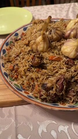 The video shows a close-up of a large, round, decorative plate filled with freshly prepared pilaf (also known as plov). The dish consists of long-grain rice, chunks of meat (likely lamb or beef), and shredded carrots. Several whole heads of garlic, some with their stems still attached, are placed on top of the pilaf as a garnish and for flavor. The plate has a colorful, patterned rim with blue, green, and red motifs.

In the background, out of focus, a light yellow plate is visible to the left, and a fork is partially seen on the right. The food is resting on a white tablecloth with a subtle, embossed or woven floral pattern. At the 0:03 mark, a hand briefly enters the frame to adjust one of the garlic bulbs on the dish. The scene appears to be an indoor setting, likely a dining table, suggesting a meal is about to be served or enjoyed. The lighting is warm and inviting. There is muffled speech in the background audio. This scene is located in Пролетарский, Russia.
