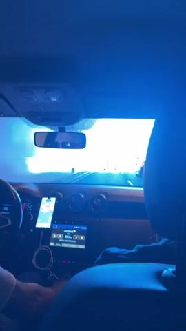 A video shows the view from the back seat of a moving car. The driver, reflected in the rearview mirror, wears sunglasses. A cell phone is mounted on the dashboard, displaying a map. The radio shows that it's playing a radio station. Outside, the car is driving down a highway, with a yellow car visible ahead in the same lane. The sky is bright and clear.