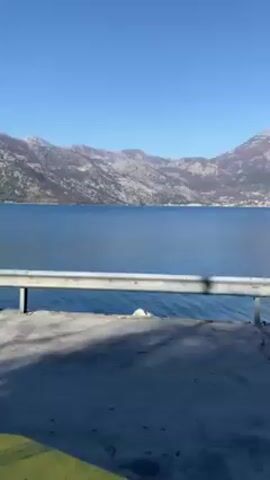 The video shows a view of a lake and mountains from a bus stop. The bus stop has a clear roof and walls, and a bench inside. The lake is blue and still, with mountains in the background. The sky is clear and blue. A guardrail is in the foreground, along the edge of the road. A red buoy can be seen in the lake.