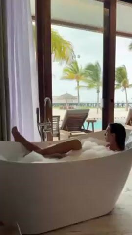 A woman is sitting in a white bathtub full of bubbles in a room with floor-to-ceiling sliding glass doors that open to a beach with palm trees and lounge chairs. She has her legs raised in the air. The room is bright and the scene appears to be from a high-end resort or vacation rental. There is a silver faucet attached to the side of the tub. The woman is facing away from the camera and has wet hair.