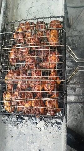 The video displays a close-up of a metal grilling basket filled with numerous pieces of meat, likely chicken, cooking over a heat source. The meat pieces are golden-brown and appear well-marinated and partially cooked, with some edges showing a darker char. They are arranged in a single layer within the grid of the grilling basket. The grilling basket itself is made of dark metal, possibly cast iron or blackened steel, and has a handle visible at the top center.

The basket rests on a rough, light gray, concrete or stone surface, which forms the edge of a grill or a makeshift barbecue setup. To the right of the basket, there are visible ashes and some dark, possibly burnt, material, indicating the presence of charcoal or wood fire beneath. The background is out of focus but appears to be a wall or a structure made of similar gray material. The lighting suggests an outdoor setting, possibly during the daytime. No specific actions are shown, but the stationary shot captures the grilling process.