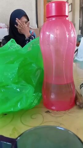 A woman wearing a hijab and a black and blue patterned shirt is sitting at a table. She is touching her face with her right hand. To her left is a crinkled bright green plastic bag. To her right is a pink plastic water bottle with a red cap. The bottle contains some liquid, with the lower half being a light brown color and the upper half being clear. The table has a yellow tablecloth with a textured pattern. In the foreground, a dark colored object, possibly glasses, is partially visible. The background is out of focus but appears to be an indoor setting with some wall decorations and furniture.