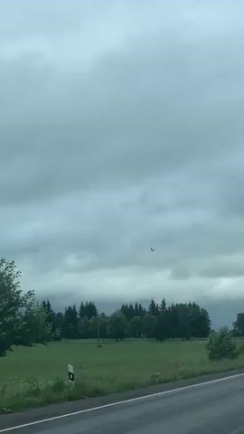 The media displays a slightly overcast sky above a rural landscape. A small airplane is visible in the upper part of the frame, flying against the cloudy backdrop. Below the sky, a dense line of evergreen trees forms a distinct horizon. In the foreground, a green field stretches out, with some scattered trees and a single wooden pole. A paved road is visible at the bottom of the frame, suggesting a roadside view. On the right side of the frame, a portion of an orange structure with a grey top is partially visible, and a blue bus stop sign with a white bus icon is mounted on a post. Text is visible on an orange sign in the lower left quadrant, reading "ДОРОГА НА ЧЕРЕМЫКИНО" (Road to Cheremykino). The overall mood is serene and quiet, typical of a rural setting, with the presence of the airplane adding a touch of dynamism. The weather appears to be cloudy, indicating it might be a cool or overcast day, possibly late afternoon or early evening given the diffused light. The setting is clearly a roadside in a rural area of Russia, near Cheremykino. There are no people visible in the media.