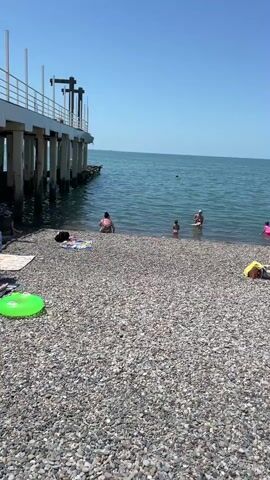 The video shows a busy pebble beach on a sunny day. Several people are in the shallow water, some swimming, others standing. On the beach, people are sunbathing on towels and lounge chairs. There are beach umbrellas providing shade. In the background, a pier extends into the blue sea under a clear sky. Some children are playing in the water.