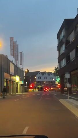 The video shows a street scene in the evening. There is a red car stopped at a railroad crossing, with the gates down and the lights flashing. Behind the car, there are a row of three connected houses. The sky is a muted blue-grey with hints of orange from the sunset. On the left side of the street are some commercial buildings with flags. On the right, a modern dark-colored building.