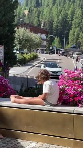 A young child with curly brown hair, wearing a white t-shirt and striped shorts, sits with their back to the camera on a golden ledge. Their bare feet are propped up on the ledge. The child is surrounded by large bushes of vibrant pink and purple flowers. In the background, a road curves uphill, lined with cars and pedestrians. Buildings, including what appears to be a store with a green cross symbol, are visible on either side of the road. The scene is set against a backdrop of a lush, green forest covering a steep hillside under a bright sky.