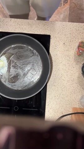 From an overhead perspective, a person standing in front of a kitchen counter pours chopped onions from a white bowl with black trim into a black skillet on a black induction cooktop. A small piece of butter sits on the side of the pan as well. The person uses a black spatula to push the onions around. The kitchen counter is light colored with a speckle pattern. A package of what appears to be pasta sits on the right side of the counter. The floor is tile.
