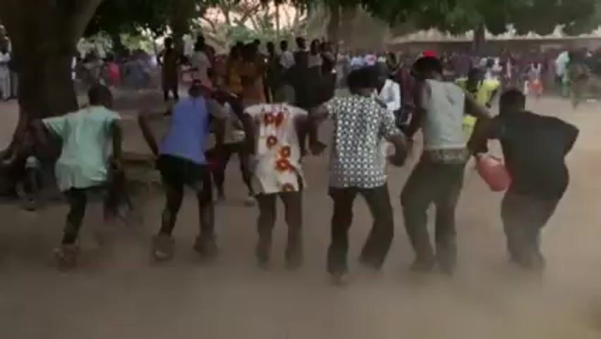 A crowd of people is gathered outdoors, kicking up dust as they dance. A person wearing a costume of long green grass moves through the crowd with a cane. Most of the people are wearing casual clothing. A large tree provides partial shade. The time of day appears to be late afternoon or early evening.