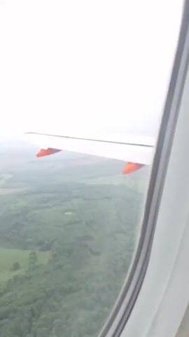 The video shows an aerial view from an airplane window, focusing on the plane's wing and the landscape below. The wing is light gray with orange details. Below, a dense green forest stretches into the distance, with some fields and a small town visible. The sky is overcast and gray. The camera appears to be stationary within the plane.