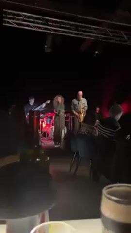 A live band performs on a stage in front of a small, seated audience in a dimly lit room. The band consists of a female vocalist, a saxophone player, a guitarist, a drummer, and someone playing a keyboard. The stage lighting is mostly red, with a metal truss visible above. In the foreground, there is a table with glasses and a partially empty bottle of wine. People are sitting in chairs facing the stage, watching the performance. The audience is mostly adults.