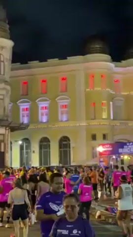 The video shows a crowd of people, mostly wearing running clothes in purple and pink, gathered at night in what appears to be a city square. There are several large, ornate buildings around the square that are brightly illuminated with spotlights. A stage or platform is decorated with signs and banners. The atmosphere seems festive, and people are moving about, some walking, some running. The video is a bit shaky and the focus is somewhat blurry at times.