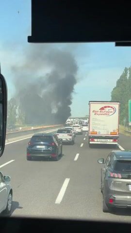 The video shows a traffic jam on a highway. The camera view is from within a vehicle. In the distance, a large plume of black smoke rises into the air on the left side of the highway, beyond a concrete barrier. Cars are lined up in multiple lanes, with a semi-truck on the right. There are some trees in the background. The sky is overcast. The car in front of the camera car is a dark blue Mazda.