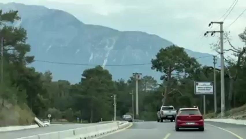 The video shows a road with cars and trees and a large mountain in the background. The mountain has a bit of snow on top. The sky is overcast. There are power lines and utility poles visible next to the road. A sign is present, though the writing on it is illegible. The video seems to be taken from inside a moving car.